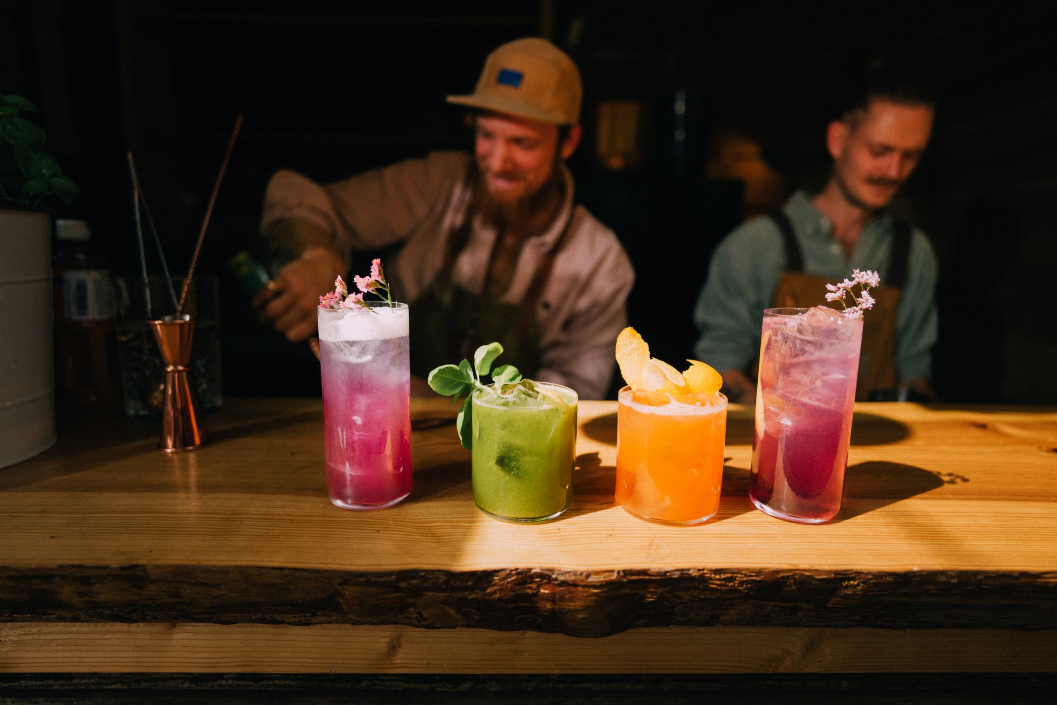 A vibrant collection of colorful cocktails displayed on a rustic wooden counter with bartenders preparing drinks in the background, creating an atmospheric and lively setting in a cozy bar. Part of a series with video